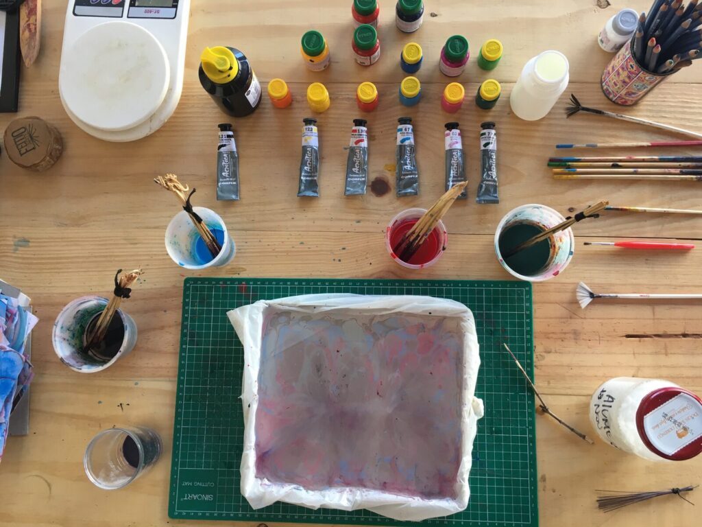 Materiais de marmorização organizados sobre a mesa do ateliê: potes e bisnagas de tinta guache e acrílica, copos com pincéis de palha e piaçava amarrados, e a cuba com gel já com um padrão marmorizado