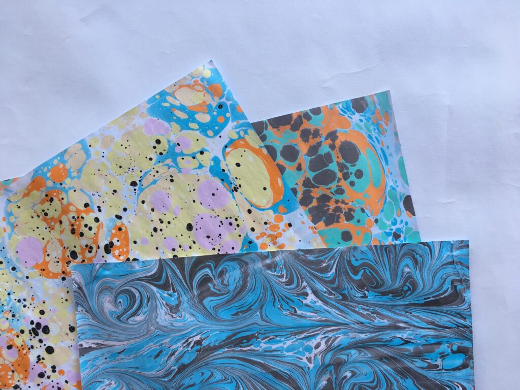 "Três folhas de papel marmorizado com tinta guache já secas, com padrões diferentes em verde e branco, rosa e azul turquesa, e laranja e azul"