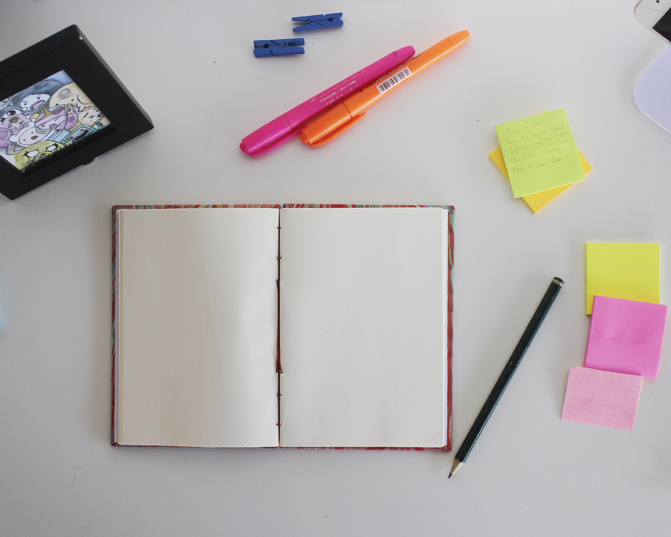 Caderno com costura copta aberto sobre uma mesa, páginas completamente planas, cercado por canetas coloridas e post-its