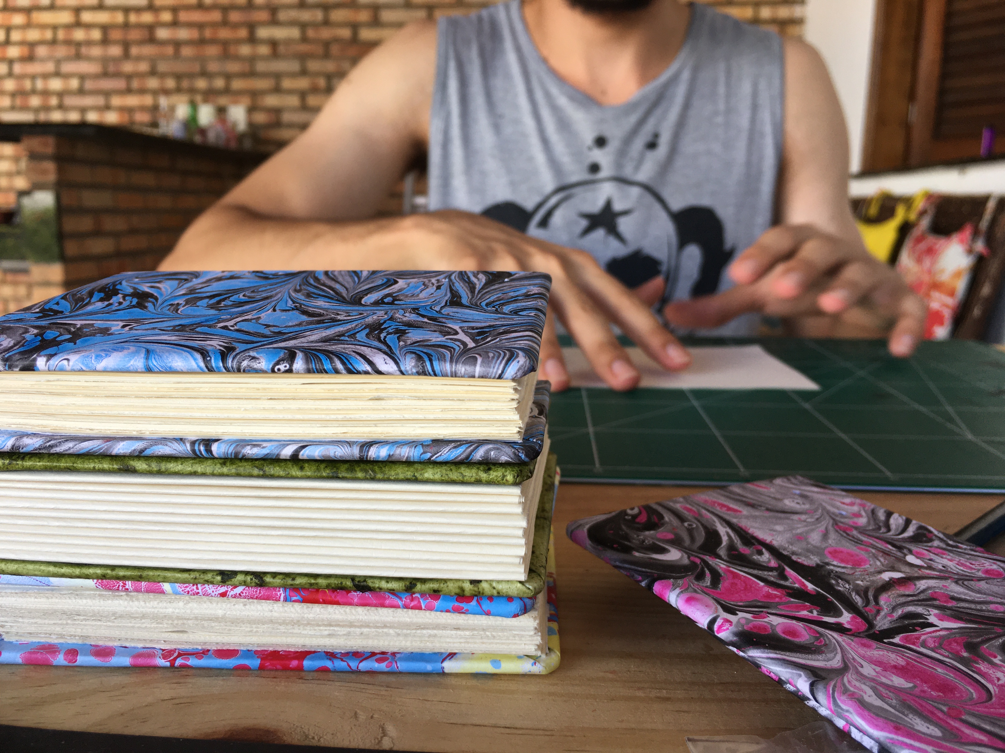 Pilha de cadernos com costura copta e capas marmorizadas coloridas em primeiro plano, com um artesão trabalhando sobre um mat de corte verde ao fundo, no ateliê da Feitio Arte