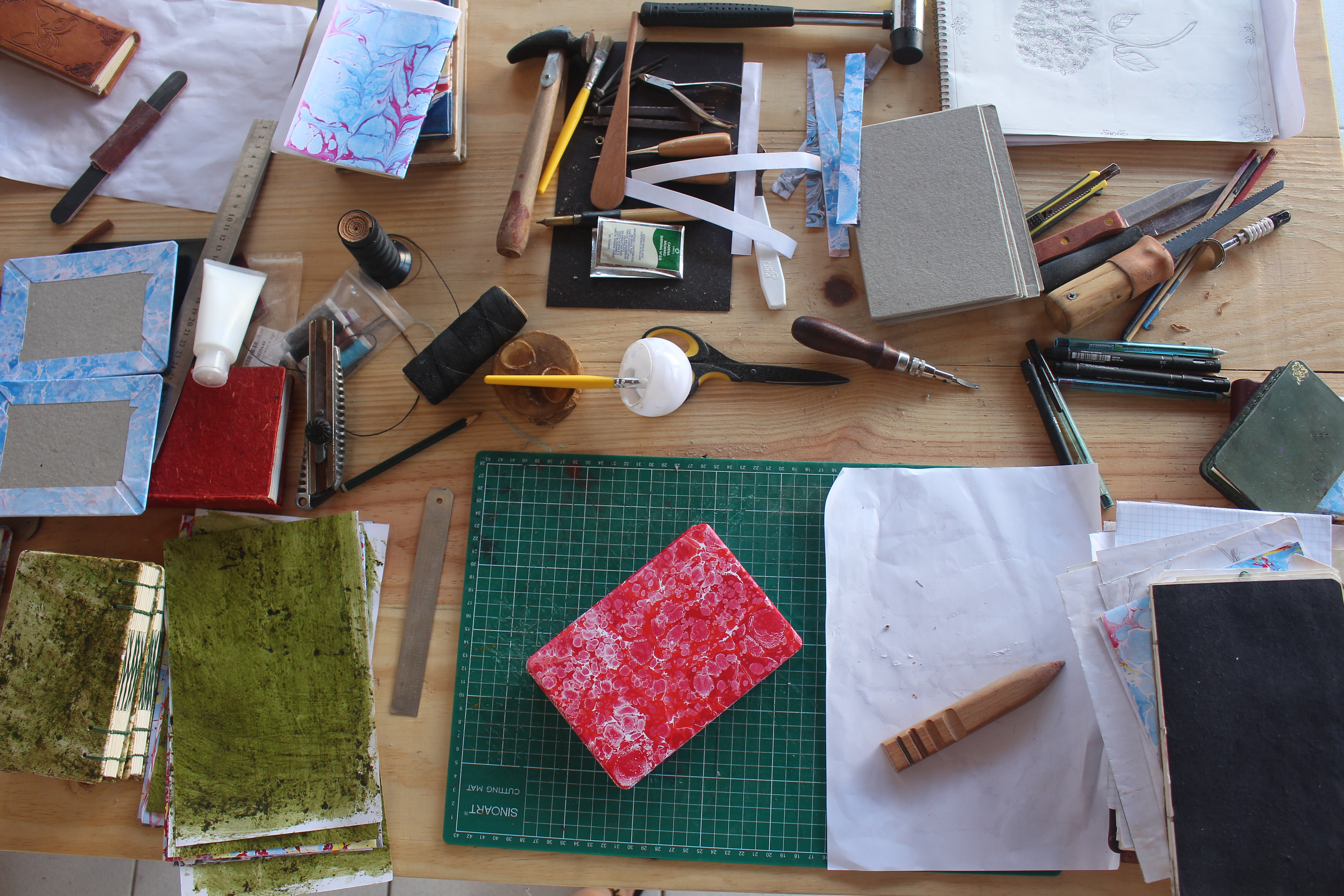 Mesa de trabalho do ateliê vista de cima, com ferramentas de encadernação espalhadas — martelo, estiletes, réguas, carretel de linha, papelão para capa, tiras de papel marmorizado e um caderno com capa marmorizada vermelha sobre um mat de corte